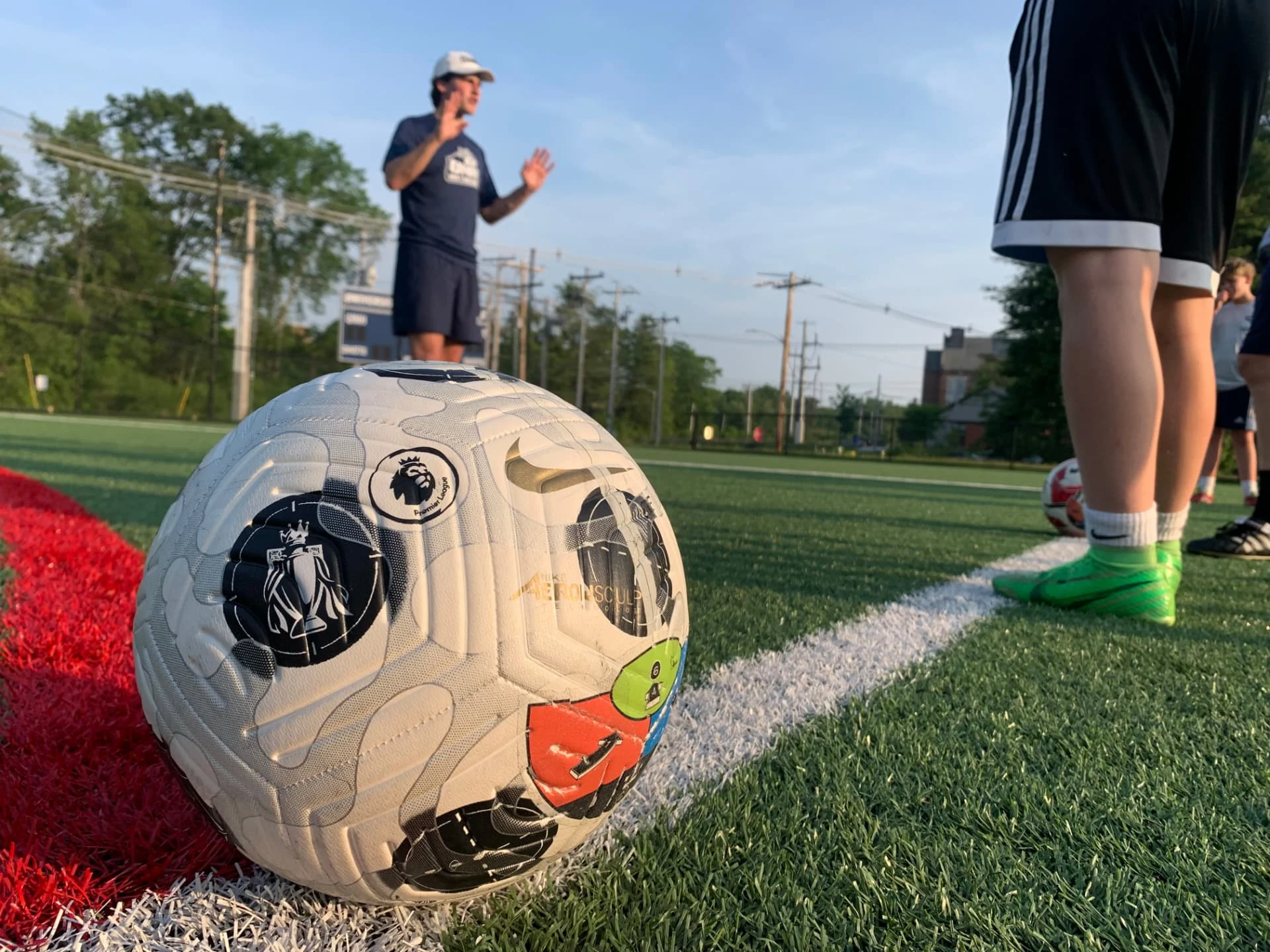 KickMap Trainer applied to a match ball on the turf during a golden-hour training session, with a coach directing play behind it.